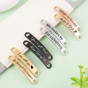 Pode incluir: Uma coleção de etiquetas de metal personalizadas em ouro, prata e preto, cada uma gravada com frases motivacionais e nomes. As etiquetas estão dispostas sobre uma superfície branca, com um fundo verde claro e folhas verdes.