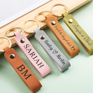 Könnte beinhalten: Eine Sammlung personalisierter Leder-Schlüsselanhänger in verschiedenen Farben, darunter Braun, Rosa und Grau. Jeder Schlüsselanhänger hat einen goldenen Ring und ist mit Namen oder Initialen graviert. Die Schlüsselanhänger liegen auf einer Holzoberfläche.