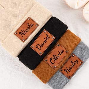 May include: Four personalized knit beanies in cream, black, brown, and gray, each with a brown leatherette patch displaying a name in black script. The beanies are folded over, and baby shoes and a wooden ring are in the background.
