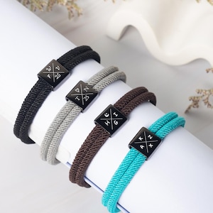 May include: Four rope bracelets in black, gray, brown, and teal, each with a black square clasp. The clasps have white symbols and letters. The bracelets are displayed on a white surface.