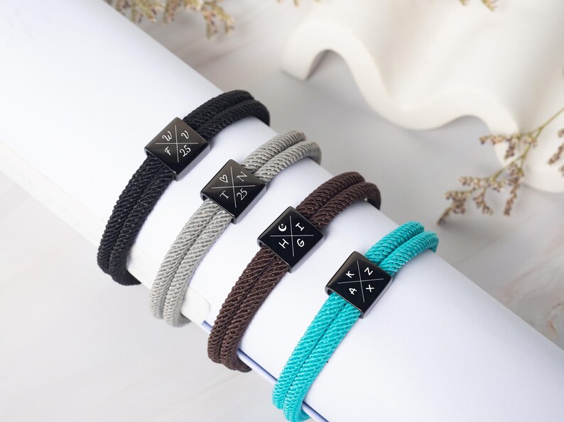 May include: Four rope bracelets in black, gray, brown, and teal, each with a black square clasp. The clasps have white symbols and letters. The bracelets are displayed on a white surface.