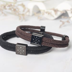 May include: Two rope bracelets, one black and one brown, with square metal clasps. The black bracelet has the letters "W", "O", "H", and "G" on its clasp. The brown bracelet has the letters "V", "L", "A", and the number "25" on its clasp.
