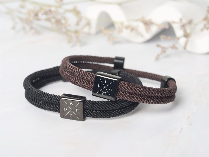 May include: Two rope bracelets, one black and one brown, with square metal clasps. The black bracelet has the letters "W", "O", "H", and "G" on its clasp. The brown bracelet has the letters "V", "L", "A", and the number "25" on its clasp.