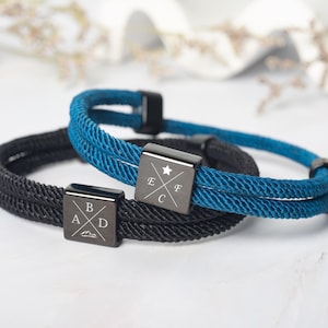 May include: Two rope bracelets, one black and one teal, with square, dark metal clasps. The clasps are engraved with letters and symbols. The bracelets are likely for everyday wear and are a stylish accessory.