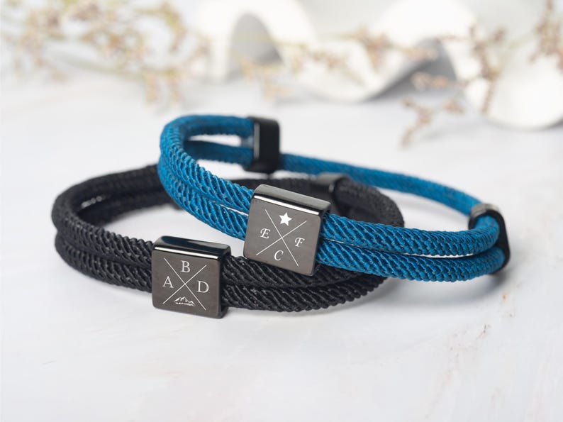 May include: Two rope bracelets, one black and one teal, with square, dark metal clasps. The clasps are engraved with letters and symbols. The bracelets are likely for everyday wear and are a stylish accessory.