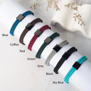 May include: A selection of colorful bracelets in blue, coffee, red, green, grey, black, and sky blue. Each bracelet features a woven cord design and a square, dark-colored clasp. The colors are labeled on the image.