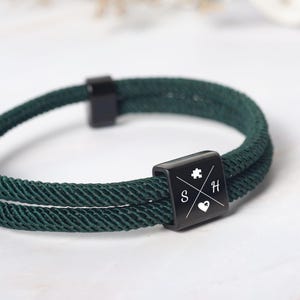 May include: Dark green rope bracelet with a black square clasp. The clasp features a puzzle piece, the letters "S" and "H", and a heart symbol. The bracelet is made of a woven rope material and has a simple, modern design.