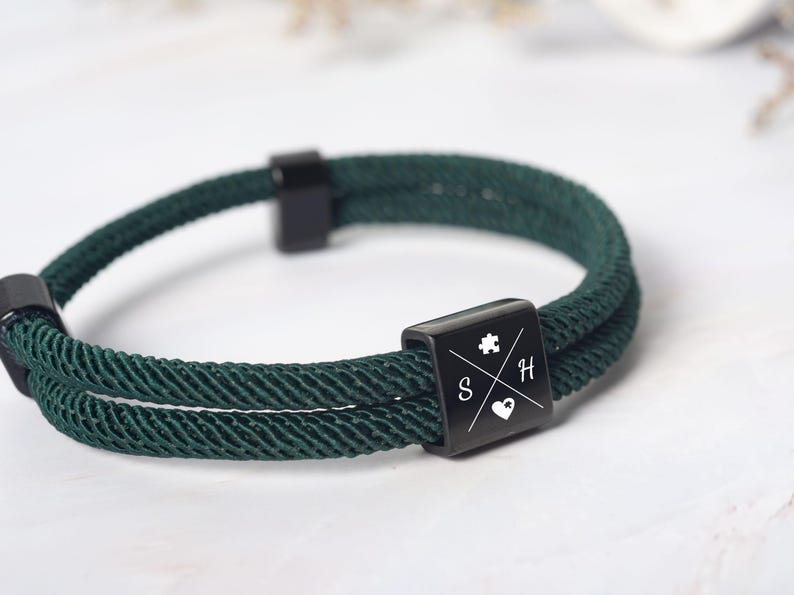 May include: Dark green rope bracelet with a black square clasp. The clasp features a puzzle piece, the letters "S" and "H", and a heart symbol. The bracelet is made of a woven rope material and has a simple, modern design.
