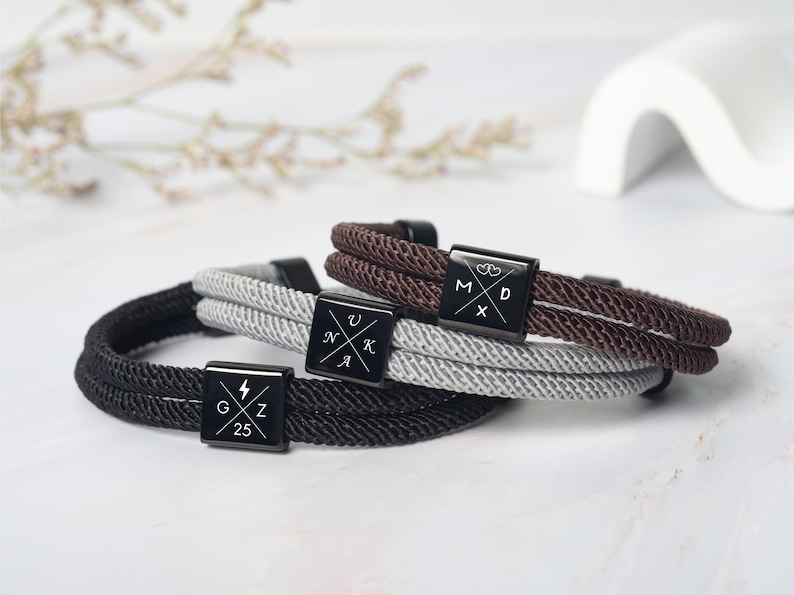 May include: Three braided rope bracelets in black, gray, and brown. Each bracelet features a black square clasp with white symbols and letters. The black bracelet has a lightning bolt and the letters G, Z, and the number 25.