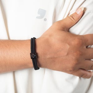 May include: Black braided bracelet with a square, black metal clasp. The bracelet is worn on a wrist, with a white shirt in the background. The clasp has a cross design.