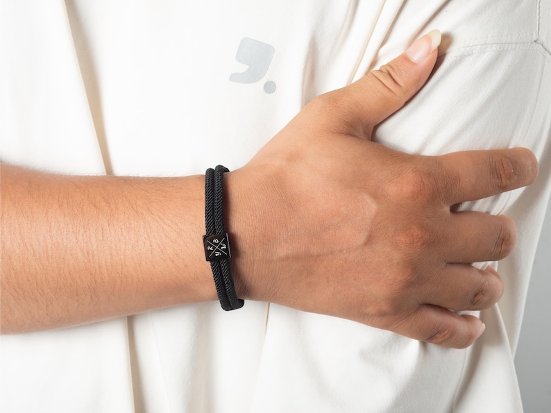 May include: Black braided bracelet with a square, black metal clasp. The bracelet is worn on a wrist, with a white shirt in the background. The clasp has a cross design.