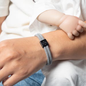 May include: A gray braided bracelet with a black square clasp featuring a white graphic design. The bracelet is worn on a wrist, with a baby's hand resting nearby. The bracelet is a stylish accessory.