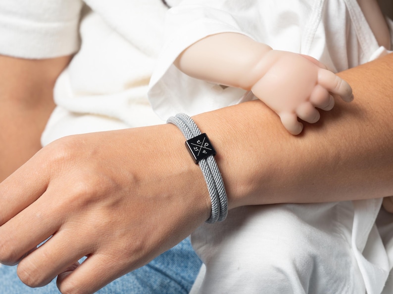 May include: A gray braided bracelet with a black square clasp featuring a white graphic design. The bracelet is worn on a wrist, with a baby's hand resting nearby. The bracelet is a stylish accessory.