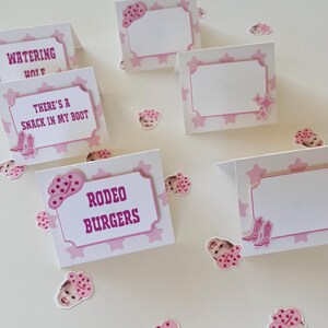Op de afbeelding: Roze en witte tafelkaarten met een roze ster- en cowboyhoedontwerp. De kaarten hebben de tekst "Watering Hole", "There's a Snack in My Boot" en "Rodeo Burgers".