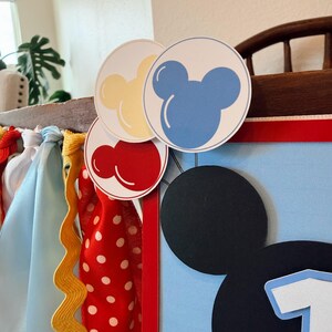 May include: A decorative banner featuring a large black mouse-ear silhouette and the number one. It is accented with red, yellow, and blue circular balloon cutouts and a variety of hanging fabric ribbons in red, yellow, white, and light blue.