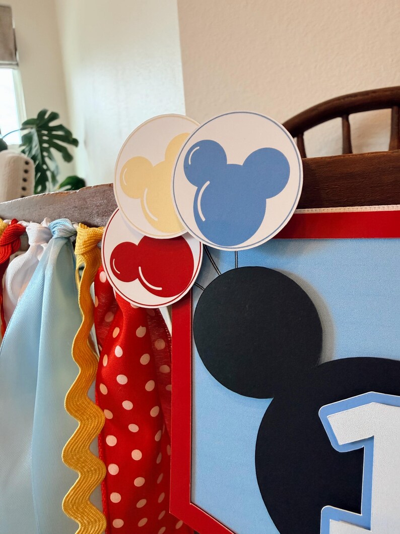 May include: A decorative banner featuring a large black mouse-ear silhouette and the number one. It is accented with red, yellow, and blue circular balloon cutouts and a variety of hanging fabric ribbons in red, yellow, white, and light blue.