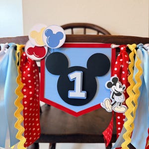 May include: A decorative banner featuring a light blue pentagonal shape with a black Mickey Mouse silhouette and the number 1 in white. It is adorned with red, yellow, and blue balloon cutouts, a Mickey Mouse figure, and red polka-dot ribbons.