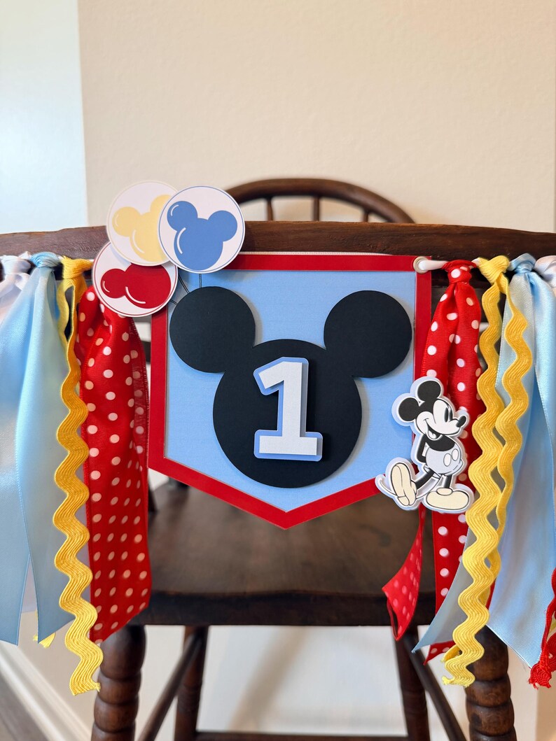 May include: A decorative banner featuring a light blue pentagonal shape with a black Mickey Mouse silhouette and the number 1 in white. It is adorned with red, yellow, and blue balloon cutouts, a Mickey Mouse figure, and red polka-dot ribbons.