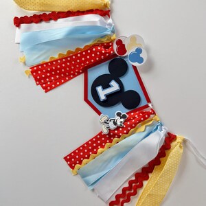 May include: A decorative banner featuring a central blue pentagonal plaque with a black Mickey Mouse silhouette and the number one. The banner is adorned with red, yellow, white, and light blue ribbons, along with small circular icons.