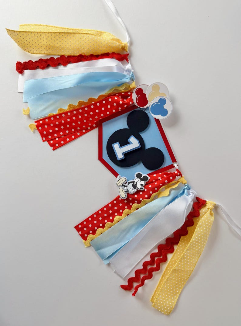 May include: A decorative banner featuring a central blue pentagonal plaque with a black Mickey Mouse silhouette and the number one. The banner is adorned with red, yellow, white, and light blue ribbons, along with small circular icons.