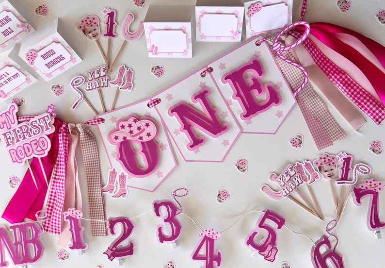 Op de afbeelding: Roze en witte eerste verjaardagversieringen met een cowgirl-thema. De versieringen omvatten een banner met de tekst "ONE", cupcake-toppers en een reeks cijfers.