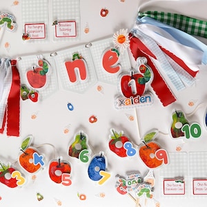 May include: A birthday party decoration set featuring a banner with the word one, colorful ribbon tassels, and cutouts of fruits and a caterpillar. The set includes cupcake toppers with the name Xaiden, numbered fruit tags, and small cards.