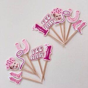 Op de afbeelding: Roze en witte cupcake-toppers met een cowboythema. De toppers bevatten een nummer &eacute;&eacute;n, een hoefijzer, een lasso, cowboylaarzen, een cowboyhoed en een kindergezicht. De tekst "Yee Haw!" is ook inbegrepen.
