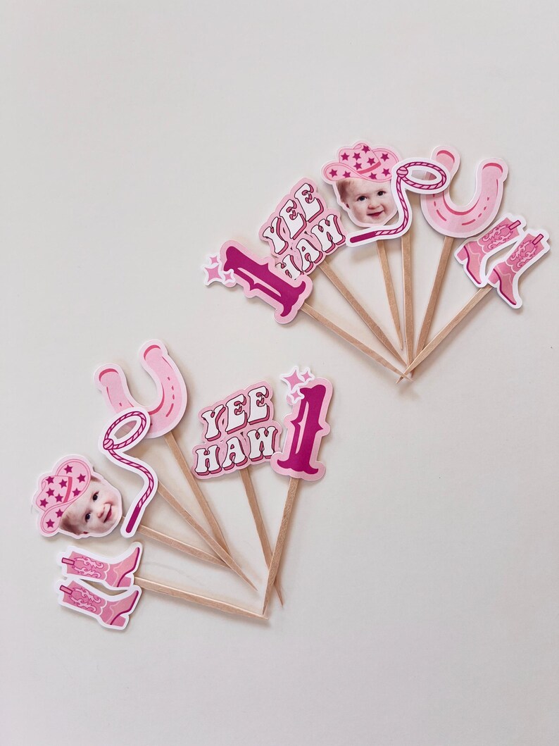 Op de afbeelding: Roze en witte cupcake-toppers met een cowboythema. De toppers bevatten een nummer &eacute;&eacute;n, een hoefijzer, een lasso, cowboylaarzen, een cowboyhoed en een kindergezicht. De tekst "Yee Haw!" is ook inbegrepen.