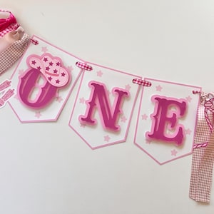 Op de afbeelding: Roze en witte banner met het woord "ONE" in roze letters. De banner heeft roze sterren en een roze cowboyhoed met een witte ster erop. De banner is versierd met roze en wit gingham lint.