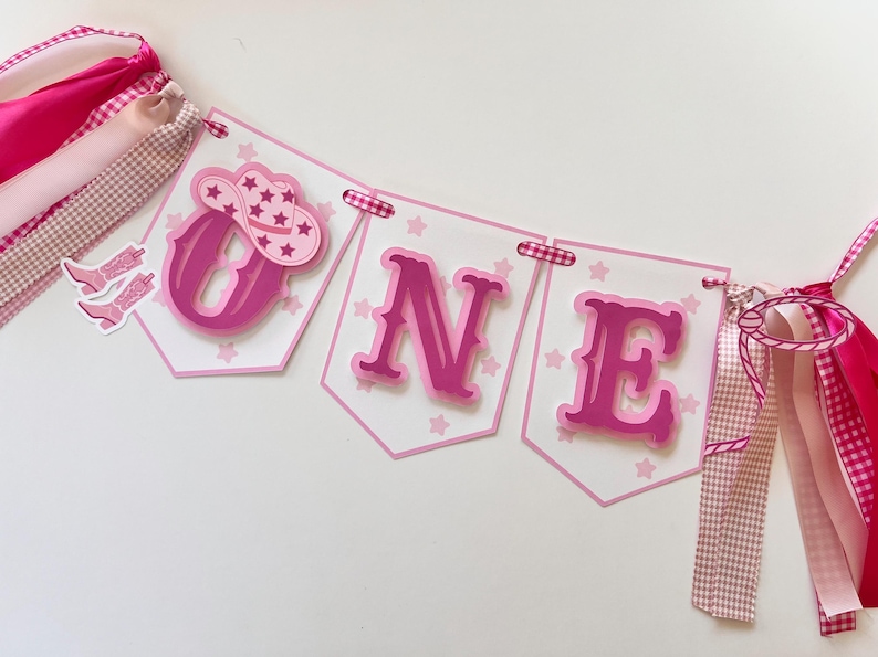Op de afbeelding: Roze en witte banner met het woord "ONE" in roze letters. De banner heeft roze sterren en een roze cowboyhoed met een witte ster erop. De banner is versierd met roze en wit gingham lint.