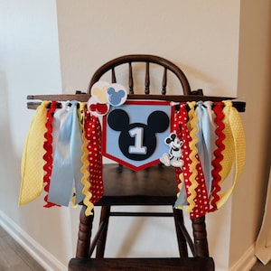 May include: A high chair banner featuring a central shield with a black mouse silhouette and the number 1, surrounded by red, yellow, and light blue ribbons. The design includes small balloon cutouts and a small standing mouse character figure.
