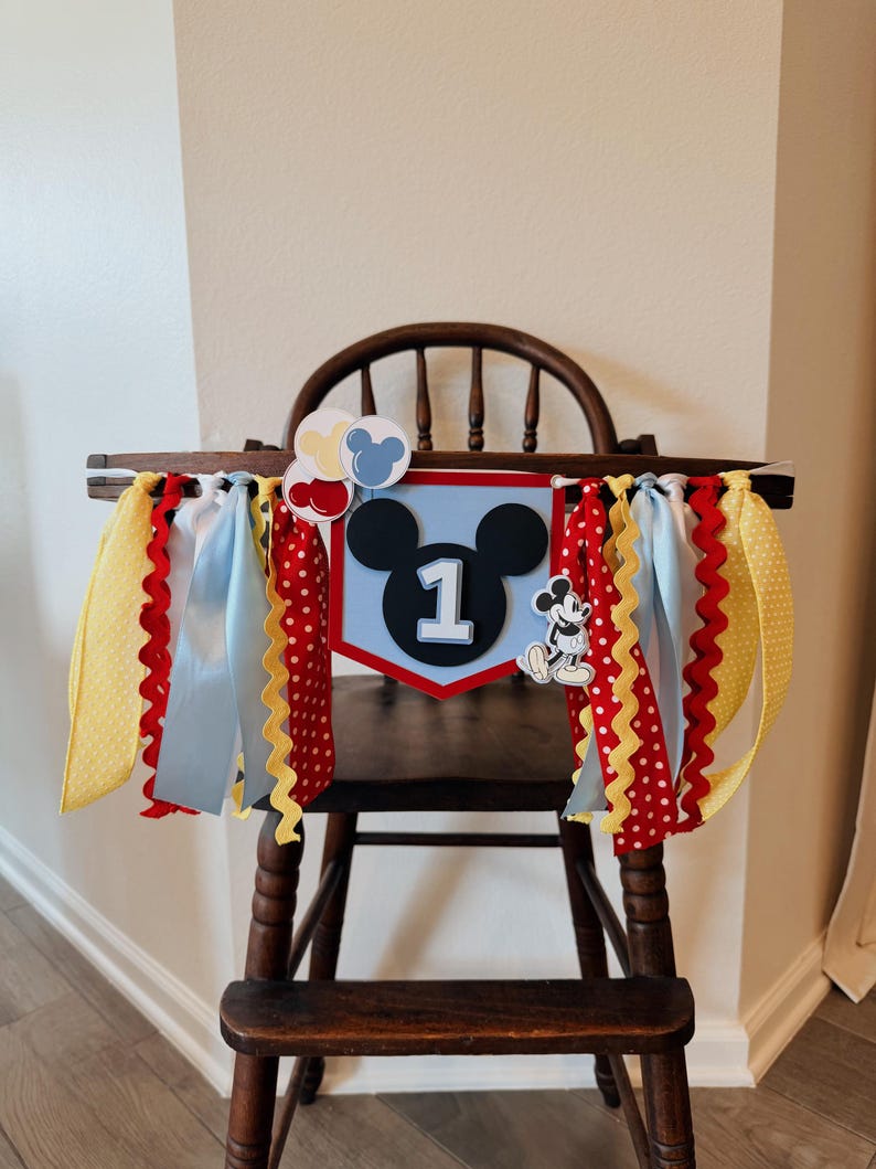 May include: A high chair banner featuring a central shield with a black mouse silhouette and the number 1, surrounded by red, yellow, and light blue ribbons. The design includes small balloon cutouts and a small standing mouse character figure.