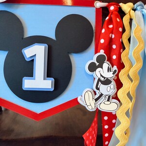 May include: A decorative party banner featuring a light blue pennant with a black Mickey Mouse silhouette and a white number one. The banner is accented with red, white, and blue ribbons, including a red ribbon with white polka dots and a yellow rickrack trim.