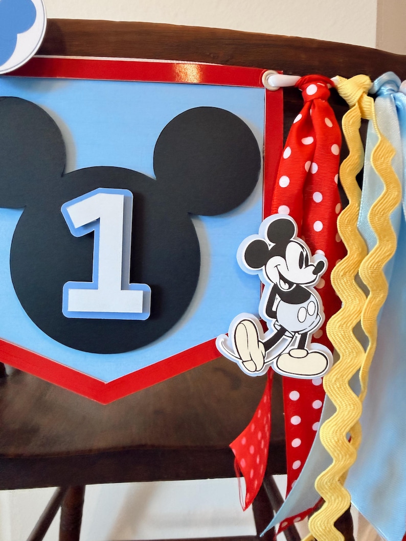 May include: A decorative party banner featuring a light blue pennant with a black Mickey Mouse silhouette and a white number one. The banner is accented with red, white, and blue ribbons, including a red ribbon with white polka dots and a yellow rickrack trim.