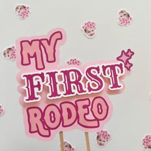 Op de afbeelding: Een roze en witte taarttopper met de tekst "My First Rodeo" in een roze en witte lettertype. De topper is versierd met kleine roze sterren.