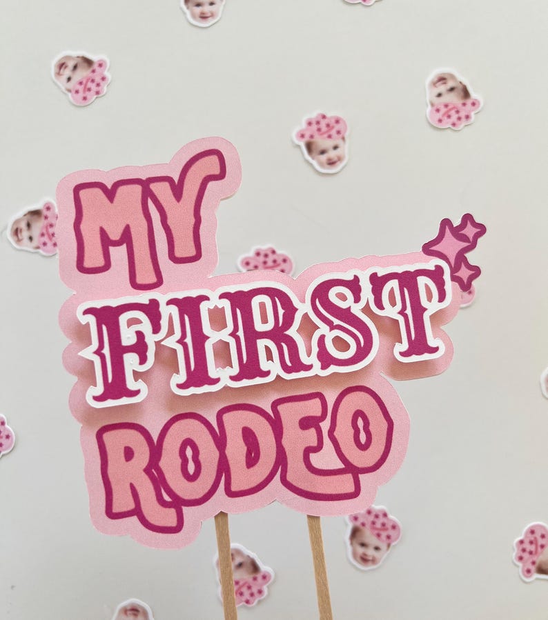 Op de afbeelding: Een roze en witte taarttopper met de tekst "My First Rodeo" in een roze en witte lettertype. De topper is versierd met kleine roze sterren.