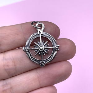Puede incluir: Encanto de brújula de plata con un diseño de estrella y las direcciones cardinales marcadas. La brújula está en una mano.
