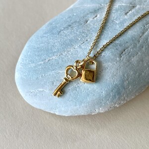 May include: A gold-colored necklace featuring a key and a padlock pendant. The key has a heart-shaped design, and the padlock is rectangular. The necklace is displayed on a light blue, textured surface.