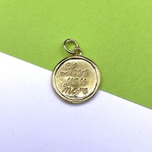 Può includere: Un ciondolo rotondo color oro con l'iscrizione "Love you more" in corsivo. Il ciondolo ha un piccolo anello nella parte superiore per il fissaggio a una catena o un braccialetto. Lo sfondo è un mix di verde e bianco.
