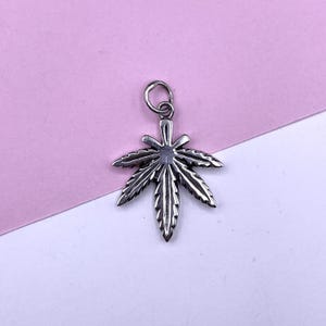 Puede incluir: Un amuleto de plata con forma de hoja de marihuana. El amuleto es detallado y tiene un bucle en la parte superior para sujetarlo a un collar o pulsera.