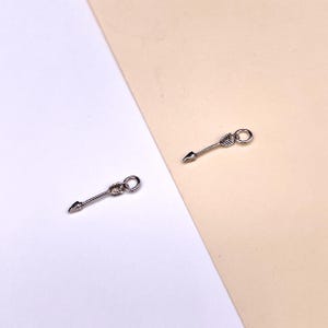 Puede incluir: Dos abalorios de flecha plateados con un bucle en la parte superior para sujetarlos a joyas. Las flechas son simples y elegantes, con una punta puntiaguda y un pequeño detalle de pluma.