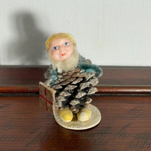 Puede incluir: Adorno navideño vintage con forma de gnomo. El cuerpo del gnomo está hecho de piñas, con una cara pintada, botas amarillas y un libro. El adorno está sobre una base blanca y tiene un cuello verde y blanco.