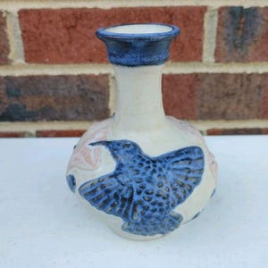 May include: A handmade ceramic vase. It features a white body, a blue rim, and a blue bird design. The bird is shown with its wings spread. The vase has a rounded base and a narrow neck. The background is a brick wall.