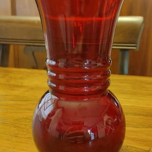 May include: A translucent ruby red glass vase featuring a bulbous base that tapers into a slender neck with three horizontal ribbed rings. The top of the vase flares outward into a wide, circular opening with a smooth, polished rim.