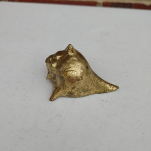 May include: A small, brass-colored seashell sculpture. The sculpture has a textured surface and a pointed top. The seashell is resting on a white surface, with a brick wall in the background.