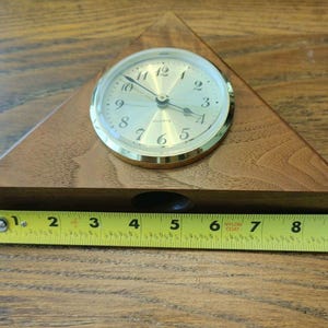 May include: A triangular wooden desk clock with a gold-toned face, black numerals, and quartz movement. A yellow measuring tape is positioned along the base, showing the clock width is approximately 9 inches from the left to the right edge.