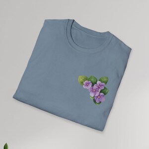 Può includere: Una maglietta azzurra con un disegno ad acquerello di viole viola e foglie verdi sul petto sinistro. La maglietta è piegata e mostrata su uno sfondo grigio chiaro. È un capo d'abbigliamento casual.