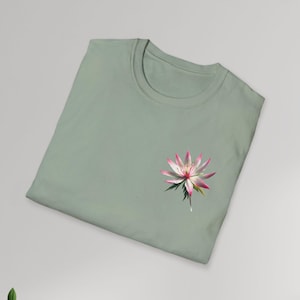 Puede incluir: Una camiseta verde salvia con un gráfico de flor rosa y blanca en el lado izquierdo del pecho.