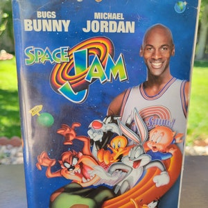 Può includere: Un nastro VHS per il film Space Jam con Bugs Bunny e Michael Jordan. Il nastro è blu con un bordo bianco e presenta un'immagine di cartone animato di Bugs Bunny, Daffy Duck, Tweety Bird e Taz. Il titolo del film è in grandi lettere bianche sul nastro.