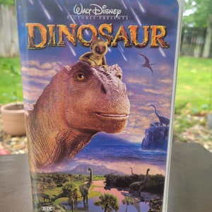 Op de afbeelding: Een VHS-cassettehoes met de Disney-film Dinosaur. De hoes toont een grote dinosaurus met een kleine lemur op zijn kop. De titel "Dinosaur" staat in grote, vetgedrukte letters. De achtergrond is een kleurrijk landschap met een blauwe lucht en wolken.